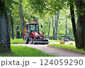 A red tractor with a trailer is parked on a gravel path in a park. Its bucket is filled with gravel. Repairing paths in a park. Preparing for the tourist season. 124059290