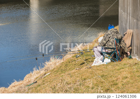 Nitra, Slovakia - 03.03.2025: An elderly man in warm clothes sits by a small overgrown river and fishes with traditional fishing rod. Nitra, Slovakia - 03.03.2025: An elderly man in warm clothes sits by a small overgrown river and fishes with traditional fishing rod. 124061306