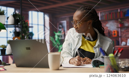 Bookkeeper in home office reviewing tax forms, reading reports on laptop and writing down information. Remotely working accountant managing finances remotely, taking notes, camera B 124063610