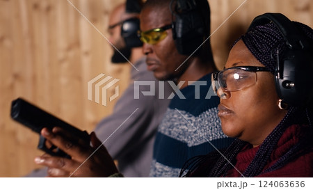 Man in firing range doing shooting sport, looking at results on paper target. Person wearing safety gear training with handgun, sharpening skills, checking points scored on bullseye, camera B Man in firing range doing shooting sport, looking at results on paper target. Person wearing safety gear training with handgun, sharpening skills, checking points scored on bullseye, camera B 124063636