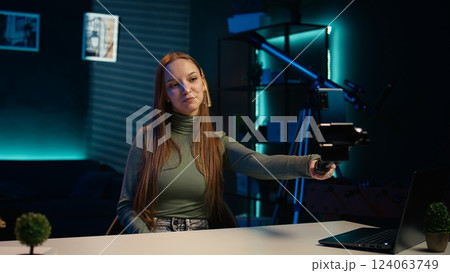 Upbeat teenager films content for online streaming channel in apartment studio using phone on selfie stick. Smiling gen z influencer at home interacts with subscribers, recording with phone, camera B 124063749
