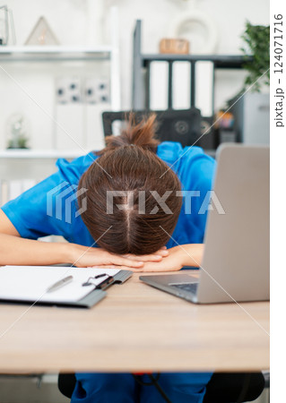 Tired healthcare professional in blue scrubs resting head on desk, surrounded by office supplies and laptop, depicting workplace fatigue. 124071716