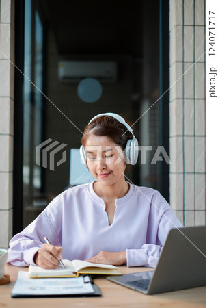 A young woman wearing headphones, studying with a laptop and notebook in a cozy home environment. 124071717