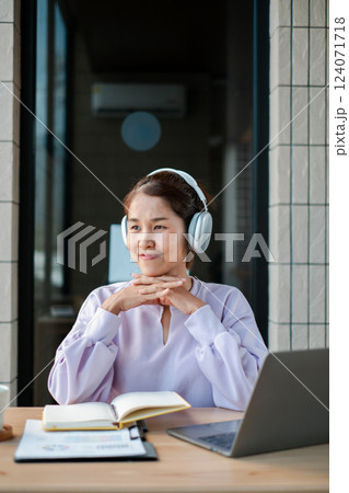 Smiling woman wearing headphones, sitting at a desk with a laptop and notebook, enjoying music while working from home. 124071718