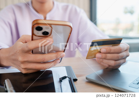 Person holding smartphone and credit card, engaging in online shopping at a home office desk with a laptop. 124071728
