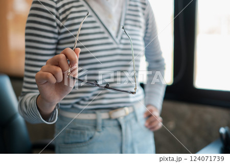 Close-up of a person holding glasses, wearing a striped shirt and jeans in a modern office environment. Close-up of a person holding glasses, wearing a striped shirt and jeans in a modern office environment. 124071739
