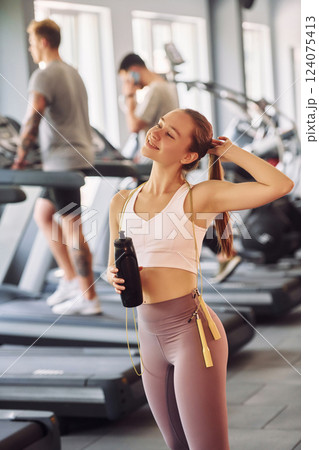 Woman in sportive clothes standing in the gym. Two men at background is running on racetrack Woman in sportive clothes standing in the gym. Two men at background is running on racetrack 124075413