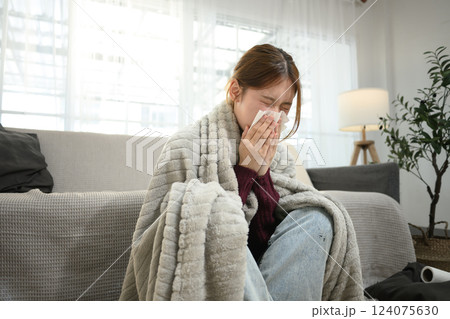 Young woman suffering from flu symptoms wrapped in blanket, sneezing into a tissue. Young woman suffering from flu symptoms wrapped in blanket, sneezing into a tissue. 124075630