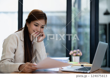 Confident business woman smiling Show successful gestures at her desk with market charts and documents in office Confident business woman smiling Show successful gestures at her desk with market charts and documents in office 124075813