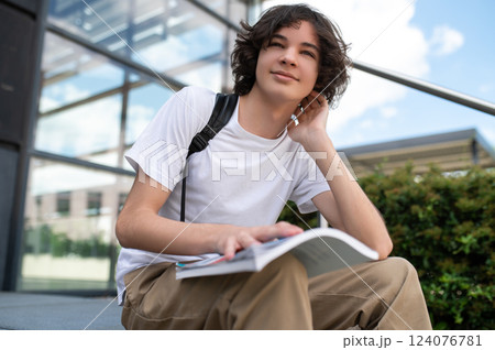 Smart guy with backpack in town embracing summer learning lifestyle holding book outdoors 124076781