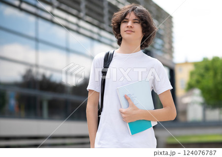 Teenager in casual outfit walking on city street carrying study bag and book 124076787
