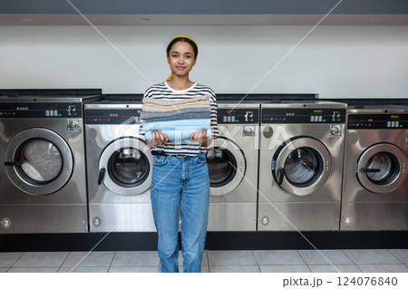 Woman in public laundromat stacking clean towels after drying them in industrial dryer 124076840