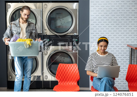 Woman in laundromat using laptop while man using washing machine for washing clothing Woman in laundromat using laptop while man using washing machine for washing clothing 124076851