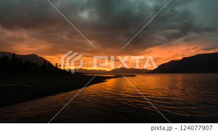 The sky burns with deep orange and gold hues as the sun sets behind distant mountains, casting a dramatic glow over the tranquil lake. Dark clouds loom overhead, adding intensity to the scene, while 124077907