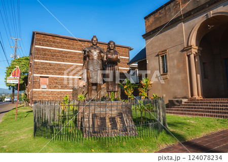 A bronze statue of a man and woman stands on a stone pedestal near a historic building with arched entrances, surrounded by tropical plants and a lawn. 124078234