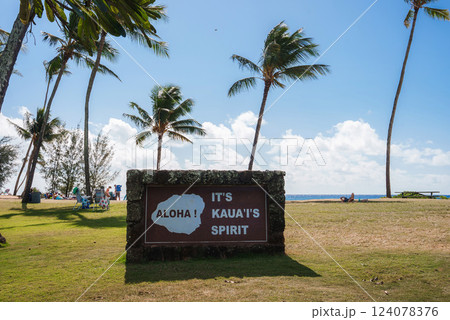 A grassy area with tall palm trees, a stone sign reading 'Aloha It's Kauai's Spirit,' and people relaxing near the ocean in Kauai, Hawaii. A grassy area with tall palm trees, a stone sign reading 'Aloha It's Kauai's Spirit,' and people relaxing near the ocean in Kauai, Hawaii. 124078376