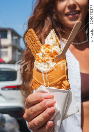 Close up of a fish shaped waffle cone filled with soft serve ice cream, caramel drizzle, a biscuit, and a wooden spoon in an outdoor urban setting. Close up of a fish shaped waffle cone filled with soft serve ice cream, caramel drizzle, a biscuit, and a wooden spoon in an outdoor urban setting. 124078377