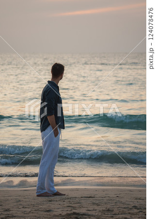 A man in white pants and a dark shirt stands barefoot on a beach in Phuket, Thailand, gazing at calm ocean waves under a serene sunset sky. 124078476