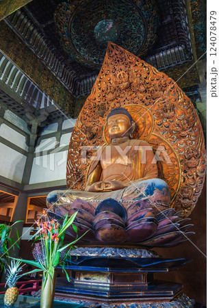 A detailed Buddha statue seated on a lotus pedestal indoors, framed by a carved halo. Tropical flowers and ornate patterns enhance the serene setting. 124078479