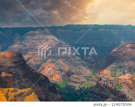 Waimea Canyon on Kauai Island features rugged cliffs with red, orange, and green layers, illuminated by golden sunlight under a partly cloudy sky. 124078504