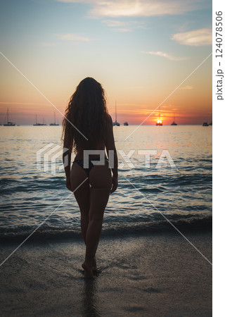 A woman with curly hair faces a vibrant sunset over calm ocean waves, with silhouetted sailboats on the horizon in Phuket or Koh Phi Phi islands. 124078506