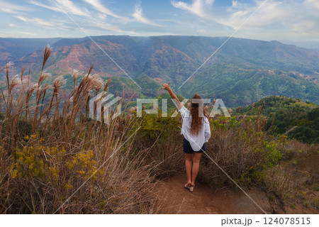 A woman stands on a dirt path surrounded by tall grass, facing Waimea Canyon's red and green cliffs under a partly cloudy sky on Kauai Island, Hawaii. 124078515