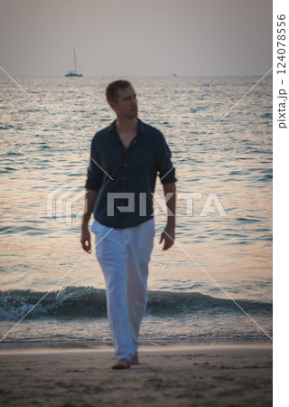 A man in a dark shirt and white pants walks along a tropical beach at sunset. Gentle waves and a sailboat on the calm sea create a serene scene. 124078556