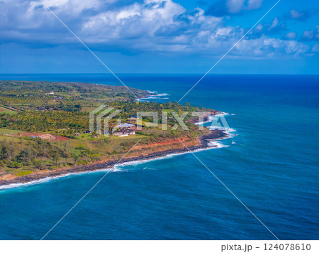 Aerial view of Kauai island's coastline in Hawaii, featuring red cliffs, lush greenery, turquoise ocean waters, scattered buildings, and a partly cloudy sky. 124078610