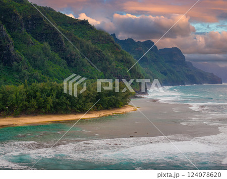 Lush green cliffs of Napali Coast on Kauai Island descend into turquoise waters. A sandy beach and warm orange pink sky complete the serene scene. 124078620