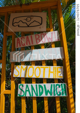 A vibrant yellow signboard displays hand painted menu items like acai bowls and smoothies, set against lush green palm fronds in a tropical setting. 124078705