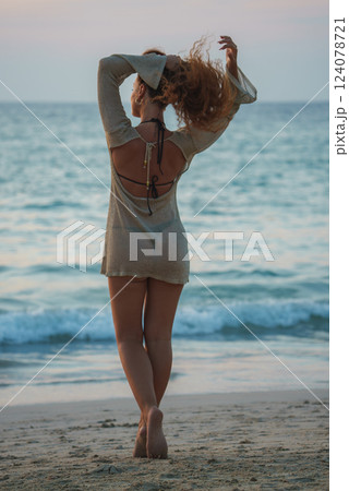 A woman in a light dress stands barefoot on a beach, facing the ocean at sunset or sunrise. Calm waves meet the shore in a serene tropical setting. 124078721