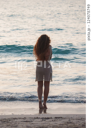 A woman with long curly hair stands barefoot on a beach in Thailand, facing calm ocean waves during sunrise or sunset, with soft light on the water. A woman with long curly hair stands barefoot on a beach in Thailand, facing calm ocean waves during sunrise or sunset, with soft light on the water. 124078749