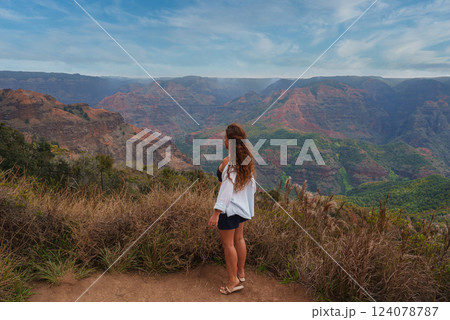 A woman with wavy hair stands on a dirt path surrounded by dry grass, gazing at Waimea Canyon's red and green cliffs under a partly cloudy sky. 124078787