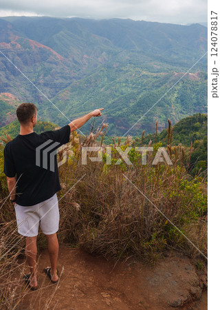 A man in a black shirt and white shorts stands on a dirt trail surrounded by dry grass, overlooking the red and green hues of Waimea Canyon under a cloudy sky. A man in a black shirt and white shorts stands on a dirt trail surrounded by dry grass, overlooking the red and green hues of Waimea Canyon under a cloudy sky. 124078817