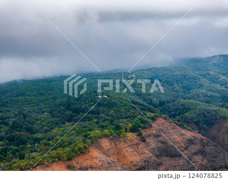 Aerial view of a lush forested ridge on Kauai island, featuring red and brown cliffs, a communication tower, and low hanging clouds creating a misty scene. 124078825