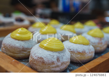 Several powdered donuts topped with bright yellow cream are arranged on a wooden tray in a bakery or market setting with a blurred background. Several powdered donuts topped with bright yellow cream are arranged on a wooden tray in a bakery or market setting with a blurred background. 124078861