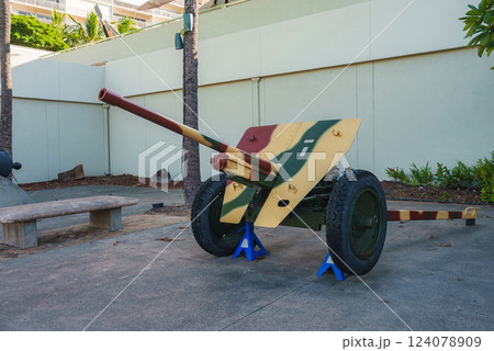 A vintage military artillery piece with camouflage paint on display outdoors, surrounded by palm trees, a concrete bench, and landscaped greenery. A vintage military artillery piece with camouflage paint on display outdoors, surrounded by palm trees, a concrete bench, and landscaped greenery. 124078909