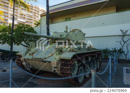 A weathered green military tank with rusted tracks is displayed outdoors near a light colored building, palm trees, and a multi story structure in Hawaii. A weathered green military tank with rusted tracks is displayed outdoors near a light colored building, palm trees, and a multi story structure in Hawaii. 124078969