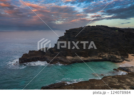 A coastal cliff of dark volcanic rock in Oahu, Hawaii, with turquoise waters, a sandy beach, and a dramatic sky transitioning from orange to blue. 124078984