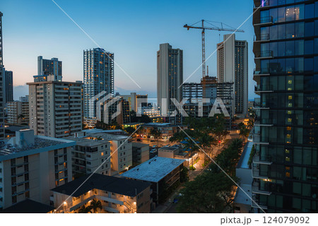 Modern high rise buildings and a construction crane in Honolulu during twilight, with warm lights, tropical trees, and the ocean faintly visible. 124079092