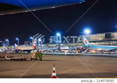 Frankfurt Airport at night with illuminated signage, parked airplanes, baggage carts, equipment, and a traffic cone on the tarmac. 124079095