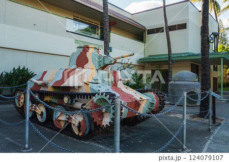 A small camouflaged tank in earthy tones displayed outdoors near the Pacific National Valor Center, surrounded by palm trees and greenery. 124079107