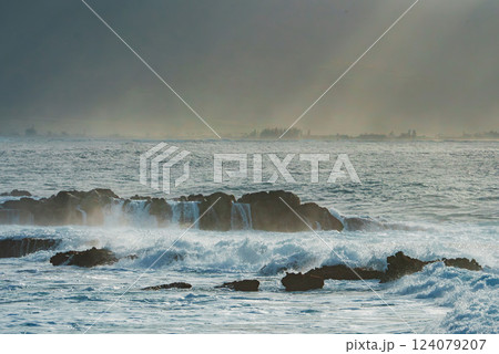 Dark volcanic rocks meet crashing ocean waves, creating white sea spray. A hazy horizon reveals distant land under soft, diffused sunlight. 124079207
