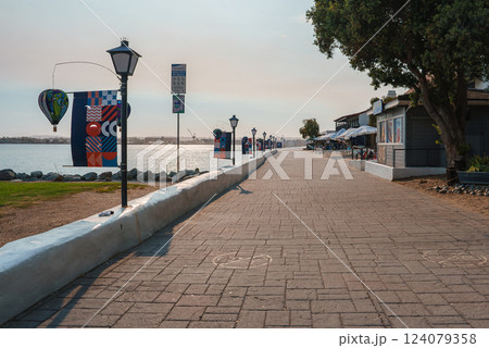 A scenic waterfront promenade features street lamps and colorful banners. Small shops with outdoor seating line the path, with calm water and distant structures visible. 124079358