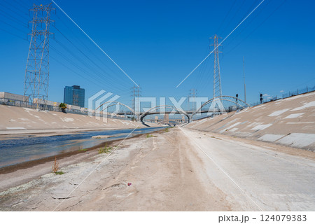 The Los Angeles River flows with concrete embankments, framed by the Sixth Street Viaduct and tall electrical towers under a clear blue sky. The Los Angeles River flows with concrete embankments, framed by the Sixth Street Viaduct and tall electrical towers under a clear blue sky. 124079383