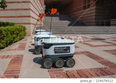 A group of delivery robots with orange flags line a brick walkway at UCLA. Steps and hedges frame the scene, showcasing technology in a university setting. A group of delivery robots with orange flags line a brick walkway at UCLA. Steps and hedges frame the scene, showcasing technology in a university setting. 124079522
