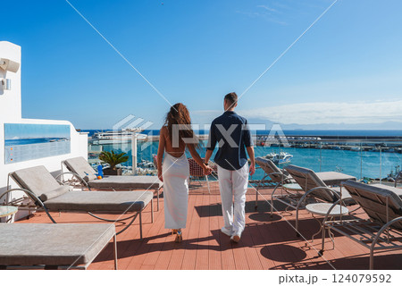 A couple walks hand in hand on a terrace with lounge chairs, overlooking a marina filled with boats. Clear blue sky and distant mountains complete the view. A couple walks hand in hand on a terrace with lounge chairs, overlooking a marina filled with boats. Clear blue sky and distant mountains complete the view. 124079592