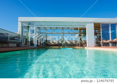 A rooftop pool area features modern Marrakech design, with sleek structures, large glass windows, seating, and lush plants under a clear blue sky. A rooftop pool area features modern Marrakech design, with sleek structures, large glass windows, seating, and lush plants under a clear blue sky. 124079785