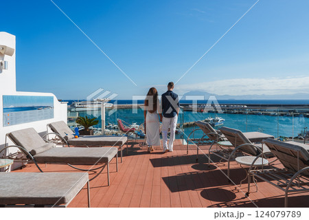 A couple stands on a terrace with sun loungers, overlooking a marina with docked boats. The scene includes a clear blue sky and distant mountains. A couple stands on a terrace with sun loungers, overlooking a marina with docked boats. The scene includes a clear blue sky and distant mountains. 124079789