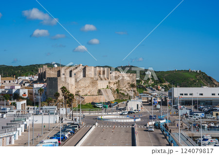 The historic Castle of Tarifa stands on a hill with stone walls and towers, overlooking modern roads and parking areas under a clear blue sky. 124079817